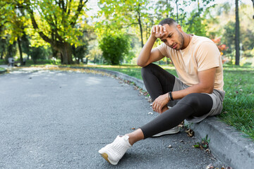 Tired african american man sitting on the ground in the park, man after jogging and doing active physical exercise overworked resting on a sunny day.