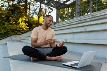 Young African American athlete fitness trainer conducts online training sitting in lotus position, man talking to physical class students using laptop and online video call.