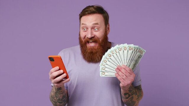 Excited happy joyful exultant young man 30s wear casual clothes use mobile cell phone hold win fan of cash money in dollar banknotes isolated on plain pastel light purple background studio portrait
