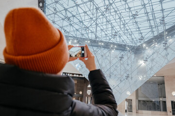 Young woman taking a photo. Inverted pyramid in the shopping mall 'Carrousel du Louvre' with people in Paris, France. Louvre museum hosts one of the biggest art collection in the world. - Powered by Adobe
