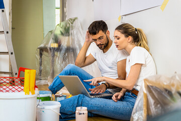 Young couple sitting on the floor choosing color via laptop for painting the wall in their home.