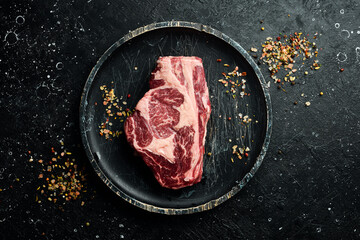 Raw Steak Ribeye Black Angus on a wooden plate ready to be cooked. On a black stone background. Rustic style.