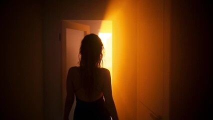 seductive woman goes to bedroom, sexy dress with open bare back black silhouette of body, walks through dark room home with evening light sunset sun rays from window. Door is open. Girl fashion model