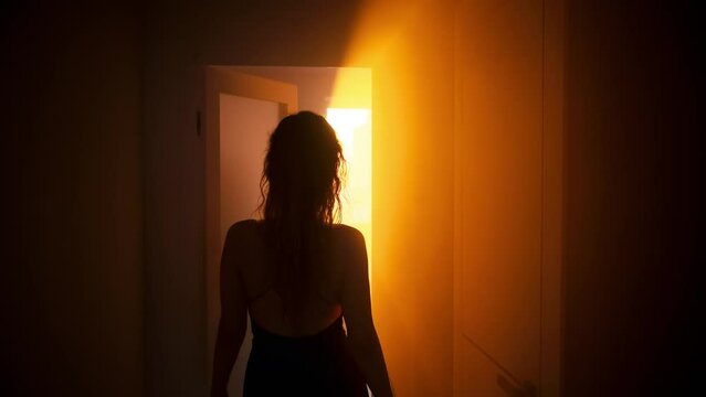 seductive woman goes to bedroom, sexy dress with open bare back black silhouette of body, walks through dark room home with evening light sunset sun rays from window. Door is open. Girl fashion model