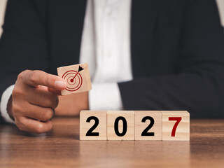 Business planning and strategy in 2027. Hand businessman holding wooden cubes with dartboard icon and wooden cubes with the letters 2027 on a table - obrazy, fototapety, plakaty