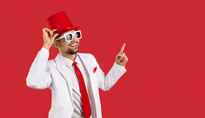 Funny man in red top hat, white suit, red tie and cool sunglasses pointing his index finger at copy space. Happy young guy in party outfit smiling and showing something on text copyspace background