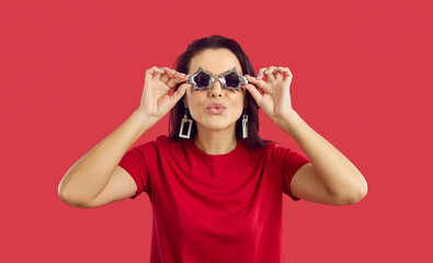 Cool and positive woman in star shape glasses makes an air kiss on vivid red background. Portrait of young woman who uses festival accessories and purses her lips for kiss while looking at camera.