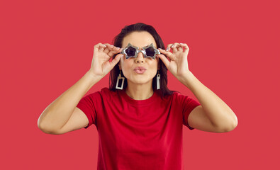 Cool and positive woman in star shape glasses makes an air kiss on vivid red background. Portrait...