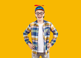 Happy preteen boy in funny cap with propeller. Portrait of cute cheerful boy wearing bifocal glasses and plaid shirt standing on yellow studio background with his hands on hips