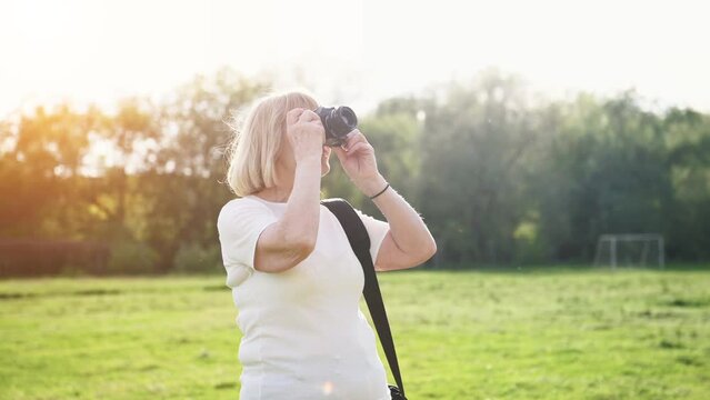Making photos by using professional camera. Cheerful senior woman is outdoors on the field at summer.