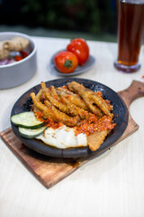Close up sambal gami ceker or fried chicken feet with sambal cooked in earthenware plate against wooden background. Served on wooden table with chili, spoon and fork as decoration