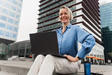 Portrait of smiling blond business woman working on laptop sitting outside office building