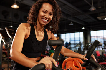 Fototapeta na wymiar African American smiling woman doing cardio workout on exercise bike in gym smiling at camera.
