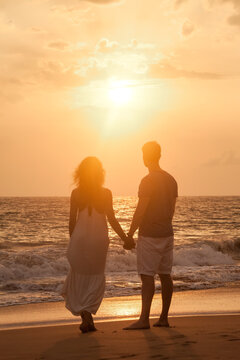 Rear View Silhouettes Lovely Couple In Love Enjoying Honeymoon Summer On Tropical Sandy Beach At Sunset Background. From Behind Happy Couple Holding Hands. Family Travel Vacation Concept. Copy Space