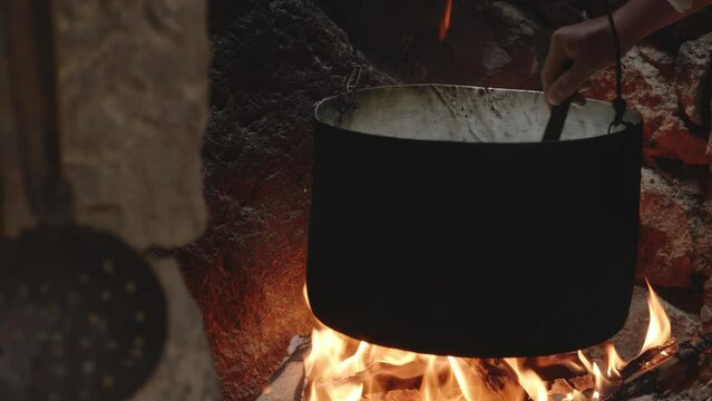 Stirring in cast iron cauldron boiling above flames in old fireplace.