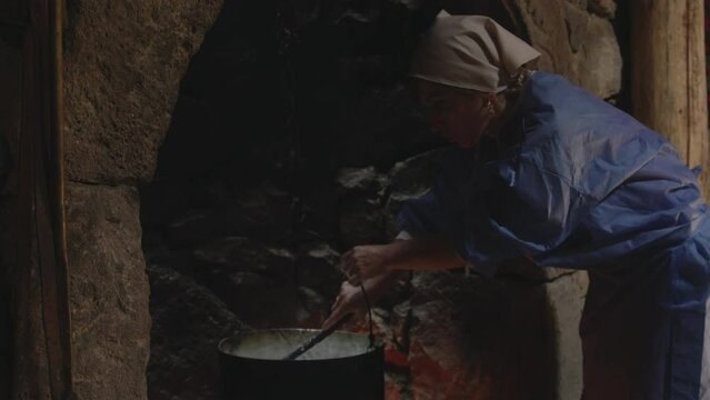 Woman in headscarf stirring milk in iron cauldron in stone fireplace.