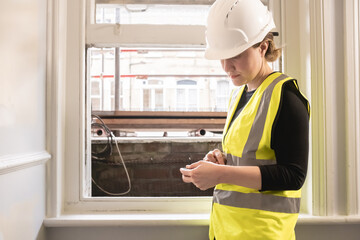 Chartered civil engineer lady using a calculator in front of a window in a construction site, wears yellow high visibility vest, ppe, personal protective equipment, site supervision, indoors