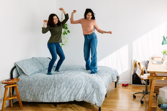 Full length view of happy young female friends in casual clothe celebrating together dancing on bed at home