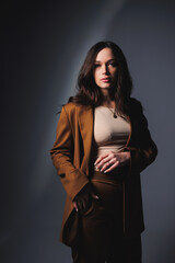 Stylish pretty young woman in brown classic suit sitting on gray background. Beautiful woman in a jacket and with makeup on her face