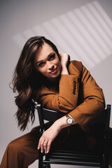 A pretty young woman with a smile on her face and wearing a brown classic suit is sitting on a chair on a gray background. Beautiful woman in a jacket and with makeup on her face