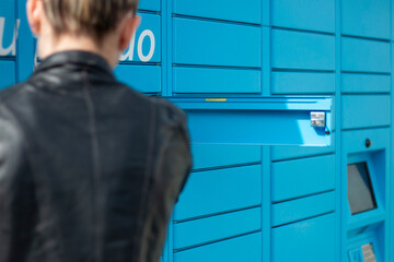 Blonde woman in a black leather jacket picking up a package in a logistics mailbox, in a blue locker on a sunny day
