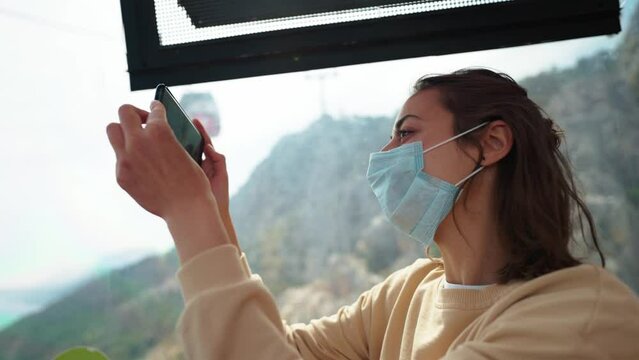 Young woman tourist in face mask sitting in cable car while moving up and filming landscape on her phone camera. cable way in Antalya, Turkey