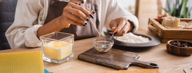 partial view of african american craftswoman dipping rope in melted beeswax near candle and cutting board, banner.