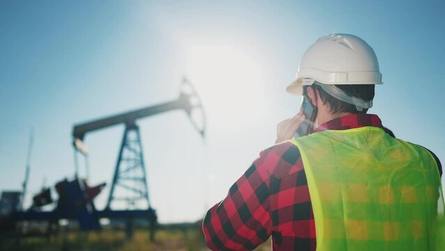 worker next to an oil rig. business oil industry concept. a worker talking on a cell phone walks with his back to an oil pump rig. engineer in a hard hat next to an oil pump lifestyle