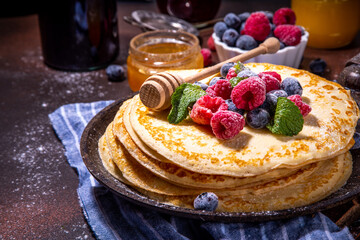 Homemade french tiny pancakes, buttercakes with berry. Stack of freshly baked thin crepes on frying pan with fresh blueberry, raspberries and honey. Healthy morning breakfast on dark background