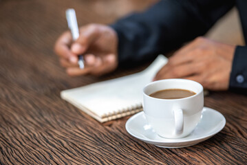 Businessman's hand taking notes message from document in mobile phone. Man write take notes by pen in notebook paper on wood table with coffee cup in park.