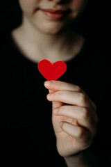 two hands hold a heart cut out of red paper - a symbol of love. A red heart made of paper on a blurry black background, part of the girl's face is out of focus.