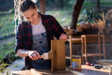 Young woman using brush applies paint or varnish on wooden board in carpentry workshop.