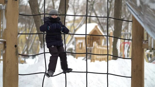The boy climbs the rope ladder. Schoolboy at a playground, climbing and holding on to rope with his hands. Playing on the playground in winter.