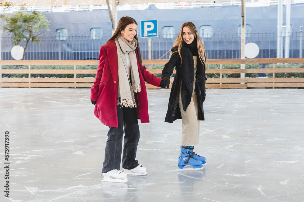 Wall mural Two pretty young girls in winter clothes holding hands laughing and skating together on an ice rink