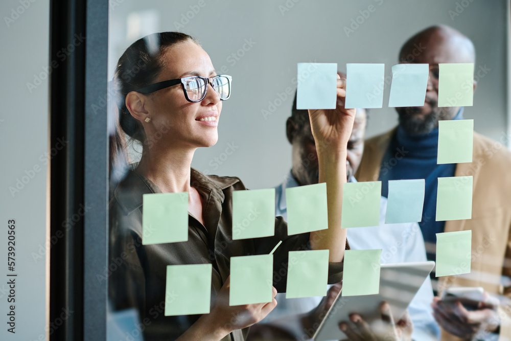 Aufkleber young female manager making notes on notepaper while standing in front of transparent board and orga