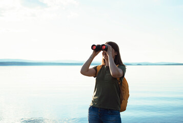 Young woman with yellow backpack looking through binoculars at birds on lake or river Birdwatching,...