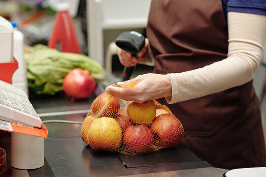 Close-up of female cashier scanning cost label on pack of red ripe apples on checkout counter in supermarket while serving customers