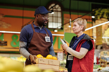 Confident mature female sales advisor with document listening to her African American male colleague carrying box of fresh oranges