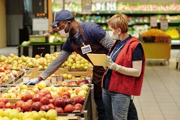 Mature manager with tablet standing by African American male colleague bending over display with fresh apples while both wearing masks