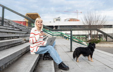 Young freelance expat woman sitting on the stairs of office building drinking coffee and working on laptop computer with her dog because it's bring-your-pets-to-work day. Web designer working outdoor