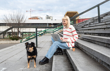 Young freelance expat woman sitting on the stairs of office building drinking coffee and working on laptop computer with her dog because it's bring-your-pets-to-work day. Web designer working outdoor