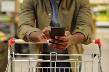 Close-up of young African American male consumer using smartphone while pushing shopping cart during his visit of supermarket © pressmaster