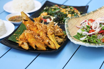 Selective focus shot of Thai food set consisting of , stir-fried liang vegetable with egg, deep-fried turmeric mullet and two plates of steamed rice on a blue table.
