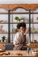 Fototapeta na wymiar smiling african american woman standing with hands in pockets of apron near natural ingredients for soap and candle making.