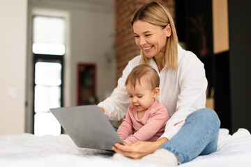 Happy mother and her little daughter using laptop computer at home, woman showing cartoons to toddler child