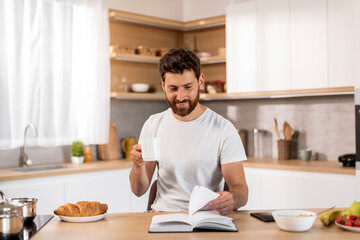 Fototapeta na wymiar Smiling adult caucasian man in white t-shirt enjoy cup of coffee, free time, reading book at table