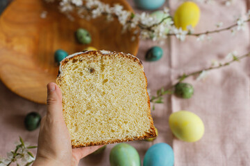 Hand holding half of baked easter cake on background of natural dyed easter eggs with spring flowers on wooden plate on rustic table.  Traditional Easter food. Happy Easter!
