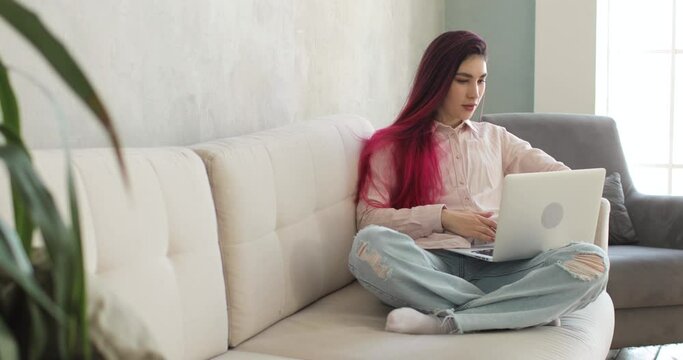 Young woman with colored hair is using laptop computer, sitting on the white sofa in the cozy living room. She is watching news on laptop and working or studing at home workplace. Home office concept.