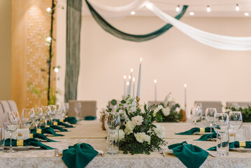 Wedding dinner table reception. A plate with a green cloth towel, knives and forks next to the plate. Flower composition with eucalyptus leaves in the center of the table and burning candles. Closeup.
