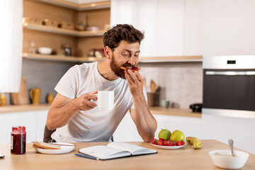 Busy handsome adult caucasian male in white t-shirt eats sandwich with jam, drinks coffee, reads book